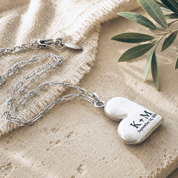 Personalized sterling silver puffed heart necklace engraved with K + M and September 14, 2024, displayed on an ivory ceramic dish with a silver chain.